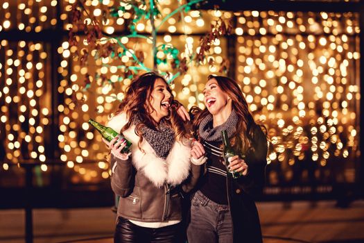 Two female friends enjoy the night out, looking each other and laughing with bottles of beer in their hands.