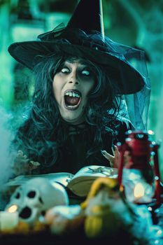 Witch with awfully face in creepy smoky green surroundings full of cobweb is reading recipe of magic drink. Halloween concept.