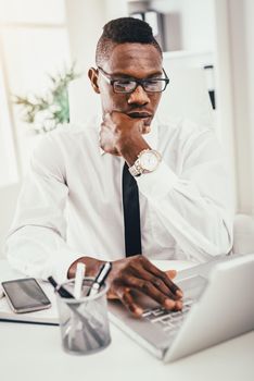 Pensive African businessman is working on laptop in modern office and planning what to do next.