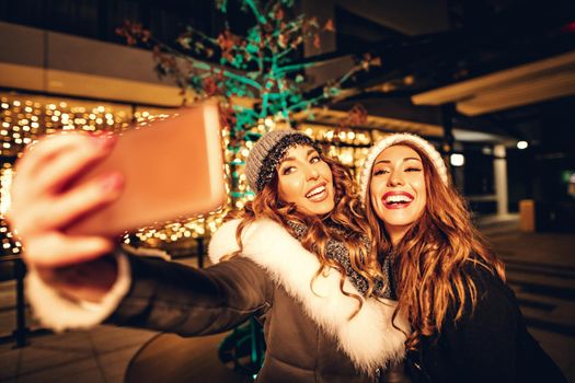 Cheerful young female friends are having fun in city street at night. They are take selfie and talking something funny.