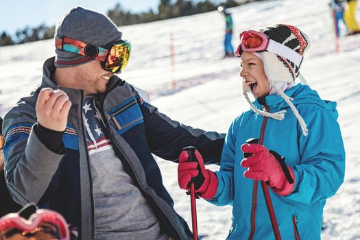 Beautiful young father and daughter enjoying in ski vacations. They are posing on sunny winter day and looking each other with smile. 