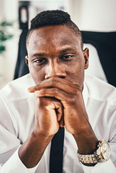 Anxious African businessman sitting in the office and holding his hands on his mouth.
