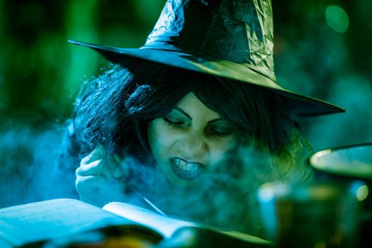 Close-up of young witch with awfully face reading recipes of magic drink in creepy surroundings and smoky background.