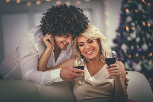 Young couple celebrating Christmas at home. They are relaxing with glass of red vine, covered with blanket on a sofa in an apartment.