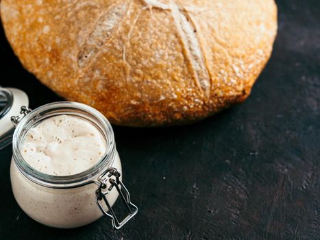 Sourdoug starter and wheat sourdough bread. Wheat sour dough starter in glass jar and delicious homemade round sourdough bread on black background, copy space. Home made sourdough bread making concept
