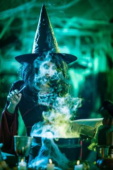 Witch with awfully face reading recipes of magic drink in creepy surroundings and smoky background.