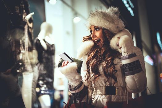 Young beautiful smiling woman having fun in the city street at Christmas time and take a photo with smartphone through the window shop.