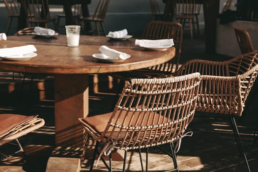 Outdoor empty coffee and restaurant terrace with wooden tables, wicker vintage chairs. Green cafe terrace on the seashore or the pedestrian street in Europe. A set table is waiting for customers