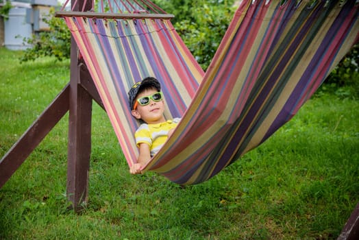 Cute little Caucasian boy relaxing and having fun in multicolored hammock in backyard or outdoor playground. Summer active leisure for kids. Child swinging on hammock. Activities for children.