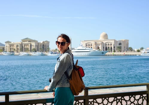 A young woman tourist with a backpack looks at the administrative region of the emirate of Sharjah with the port and ships