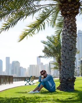 Happy tourist relaxing on lawn under palm tree in Sharjah while looking at camera. Vacation and sightseeing concept.