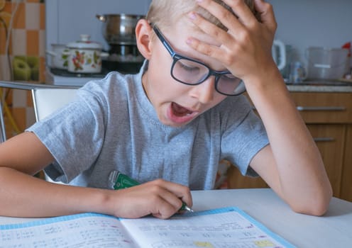 The boy solves math, does homework in the kitchen and yawns from boring tasks. The child writes the answer to the solution of the example in a notebook. The kid sits at the table and performs the task