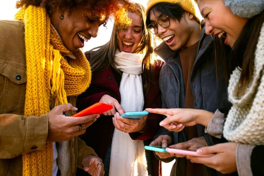 Cheerful multiracial friends looking at mobile phone together on winter day. Young woman pointing at device screen. Technology and friendship concept.