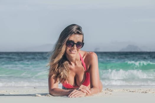 Glamorous long haired young woman in bikini and sunglasses lying on tropical beach, copy space for content