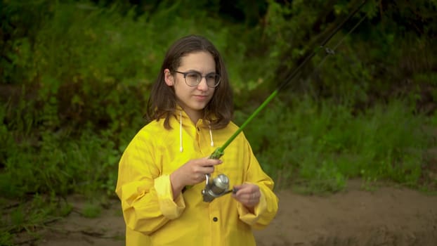 A young woman is fishing on the river bank. A girl in a yellow raincoat with a hood and glasses casts a spinning rod. Close-up. 4k