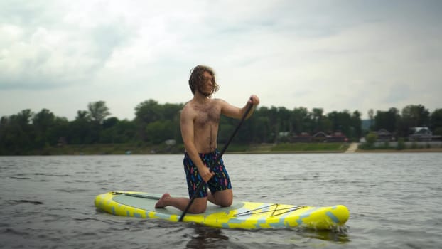 A young man swims on a sapsurf on his knees. Stylish guy with long curly hair is paddling while riding on a board. 4k