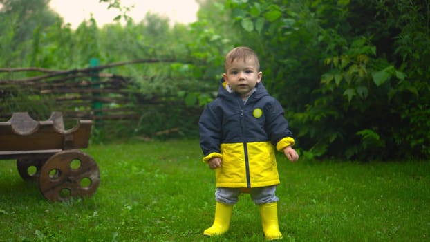 A little boy walks in the garden after the rain. The child smiles as he walks on the grass. Boy in jacket and boots. 4k