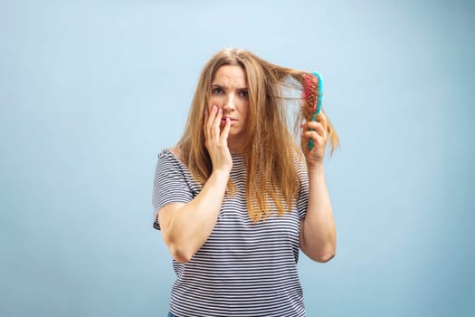 Very sad and upset young woman looking with shock at her damaged hair on blue background.