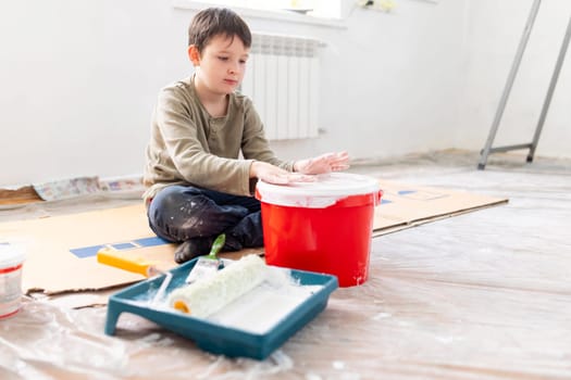 A happy child has fun sitting on the floor and banging his hands on a bucket of paint. Repair is easy. Repair in the apartment