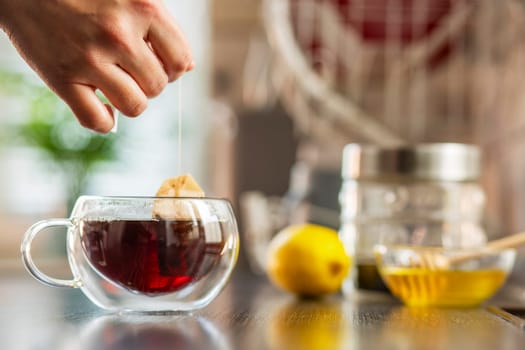 Womans hand puts the tea bag in the glass cup with hot water. Brewing a bag of black tea in a glass cup