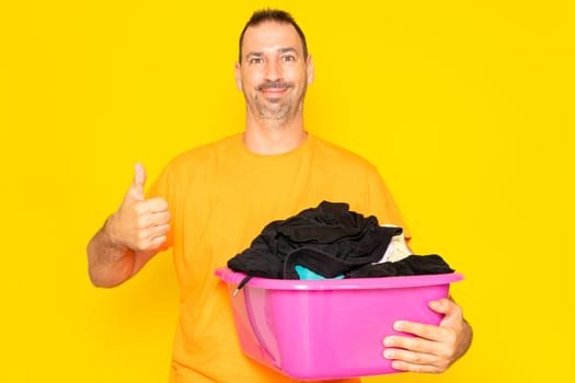 Bearded hispanic man doing housework holding plastic basket with clothes raising thumb up, excellent symbol. Isolated on yellow background