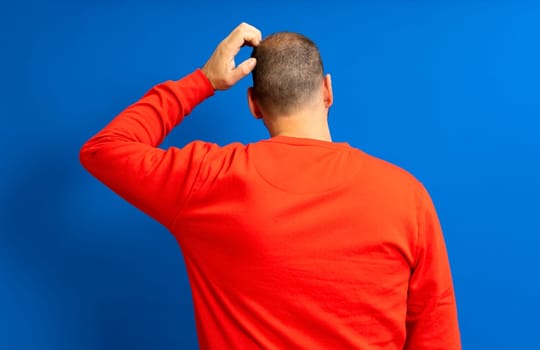 Back view of thoughtful man scratching his head over blue background. He wonders why he doesn't have luck in life and if he will always be like this