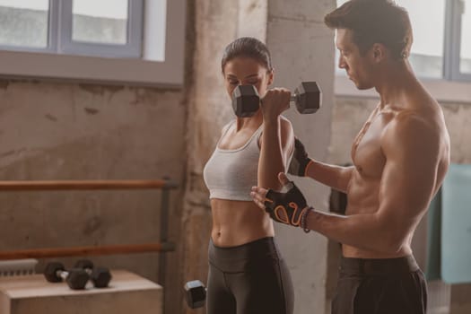 Slender lady holding dumbbells, sporty strong man helping her to correctly perform exercise for arms in the fitness club