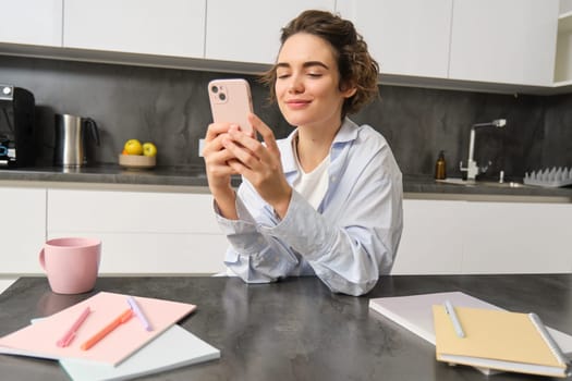 Indoor shot of beautiful woman chats on mobile phone, texts message on smartphone, orders takeaway, groceries delivery in app.