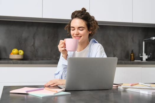 Smiling girl student, drinks coffee, studies online course from her laptop at home. Woman does her homework, works on remote, watches streaming show in internet.