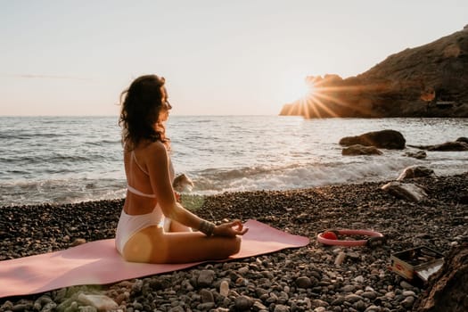 Young woman in swimsuit with long hair practicing stretching outdoors on yoga mat by the sea on a sunny day. Women's yoga fitness pilates routine. Healthy lifestyle, harmony and meditation concept.