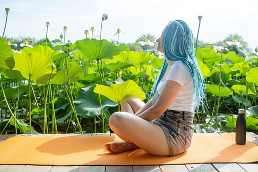 young beautiful woman with blue afro locks resting on yoga mat on wooden pierce on lotus lake enjoying nature