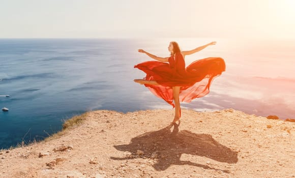 Side view a Young beautiful sensual woman in a red long dress posing on a rock high above the sea during sunrise. Girl on the nature on blue sky background. Fashion photo.