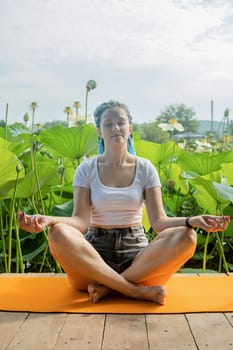 young beautiful woman with blue afro locks resting on yoga mat in lotus pose on wooden pierce on lotus lake enjoying nature