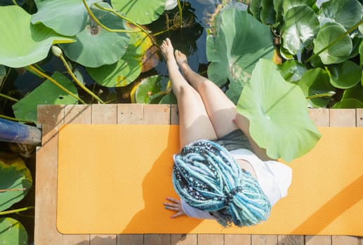 young beautiful woman with blue afro locks resting on yoga mat on wooden pierce on lotus lake enjoying nature legs in water, top view