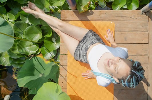 young beautiful woman with blue afro locks resting on yoga mat on wooden pierce on lotus lake enjoying nature, legs in water, top view