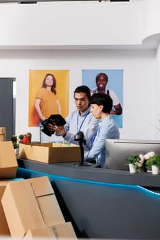 Store workers putting trendy clothes in cardboard boxes, preparing packages for delivery in modern boutique. Diverse team discussing customers orders, checking shipping report on computer