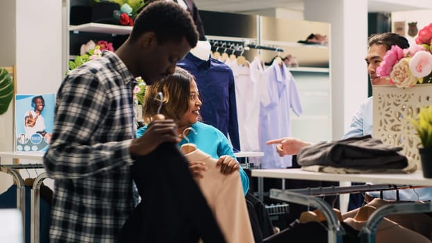 African american couple being helped by store manager, explaining clothes material during shopping session in modern boutique. Cheeful customers shopping for fashionable merchandise
