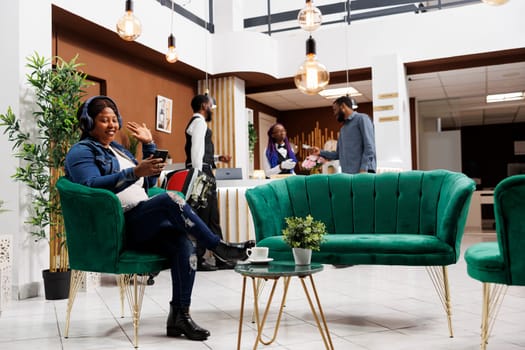 Happy woman traveler waving hand hello at smartphone camera, talking to family or friends on video call while traveling, sitting in hotel lobby. Internet communication during travel