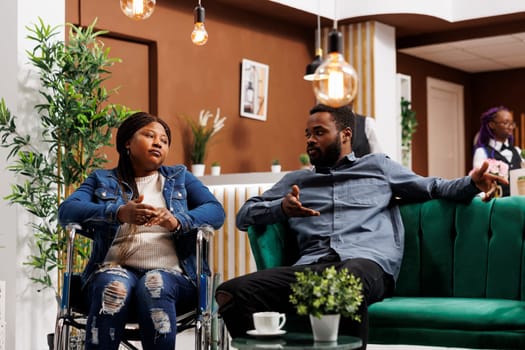 Young African American man and his disabled wife sitting in hotel lobby and talking, unhappy tired woman in wheelchair traveling with travel companion. Vacation for people with disabilities