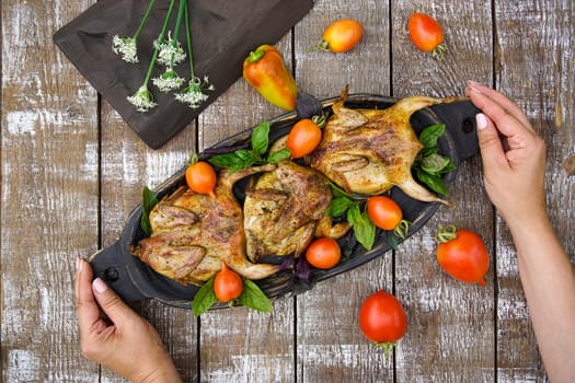 Women's hands hold a wooden dish with grilled quails. Tomatoes and bell peppers are lying on a wooden table.