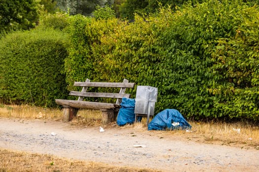 Trash and garbage in plastic bags next to a park bench with trash can at a dirt road in nature