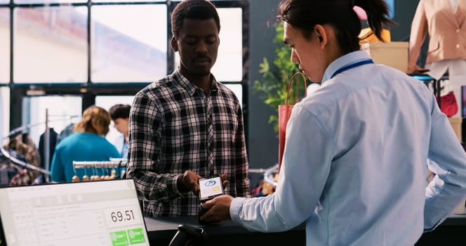 African american client buying stylish clothes, paying for trendy clothes using mobile phone nfc on pos terminal in modern boutique. Shopper man shopping for elegant merchandise. Fashion concept