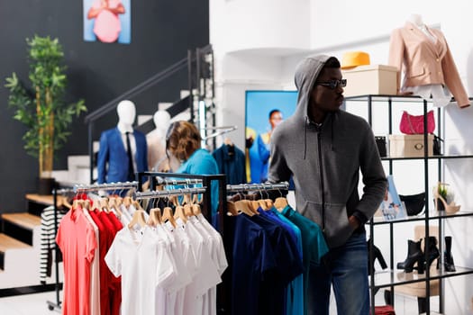 African american man with hood and glasses, trying to steal fashionable merchandise from clothing store. Robber looking around to see if someone watching him while stealing trendy clothes