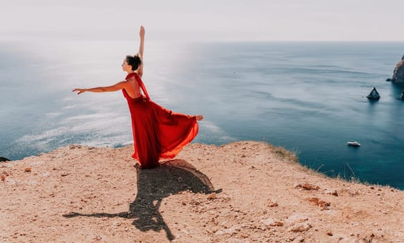 Side view a Young beautiful sensual woman in a red long dress posing on a rock high above the sea during sunrise. Girl on the nature on blue sky background. Fashion photo.