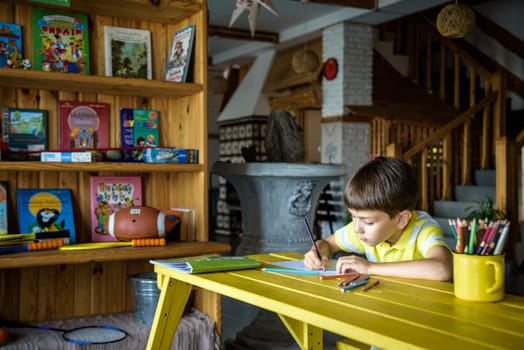 Little cutie curly haired baby boy sitting on floor of eco living room interior next to books shelf and drawing with colorful felt tip pens in album. Creative hobby for kids and playtime activity.