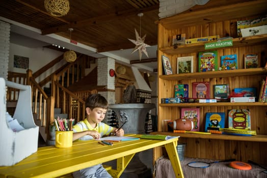 Little cutie curly haired baby boy sitting on floor of eco living room interior next to books shelf and drawing with colorful felt tip pens in album. Creative hobby for kids and playtime activity.