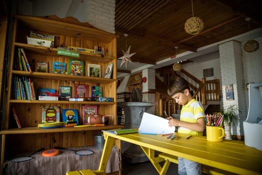 Little cutie curly haired baby boy sitting on floor of eco living room interior next to books shelf and drawing with colorful felt tip pens in album. Creative hobby for kids and playtime activity.