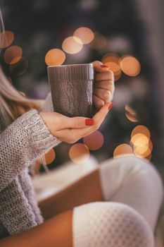 Close-up of a cute young woman sitting in the bed and holding cup of tea or coffee, surrounded with Christmas lights. 