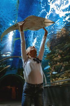 Young girl standing outstretched against aquarium glass fascinated by ocean world and touches the   devilfish in an oceanarium tunnel.
