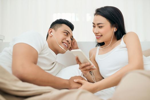 Indoor photo of loving couple lying on white sheets with smartphone.